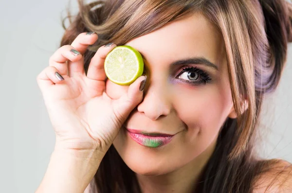 Headshot brunette, dark mystique look and green lipstick, covering one eye with open lime, looking into camera