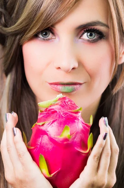 Headshot brunette, dark mystique look and green lipstick, holding up pink pitaya fruit with both hands facing camera