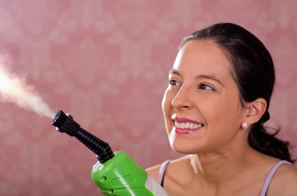 Brunette woman holding steam cleaner machine and vapor coming out, smiling to camera, pink background