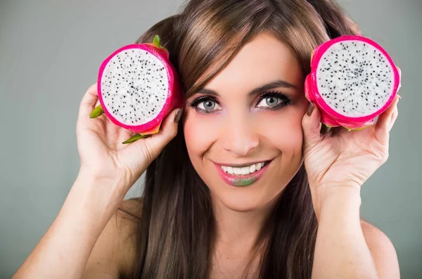 Headshot brunette, dark mystique look and green lipstick, holding up two open halfs of pink pitaya fruit with both hands facing camera