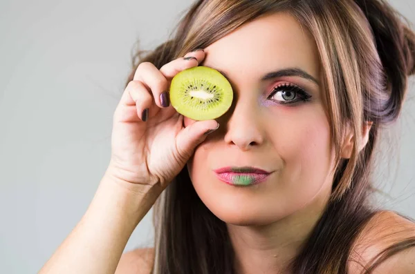 Headshot brunette, dark mystique look and green lipstick, covering one eye with open kiwi, looking into camera