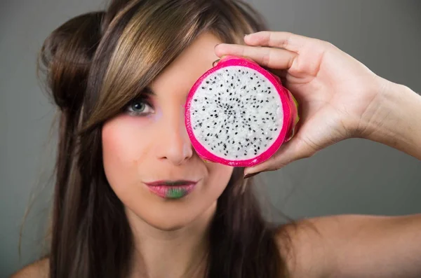 Headshot brunette, dark mystique look and green lipstick, covering one eye with open pitaya fruit, looking into camera