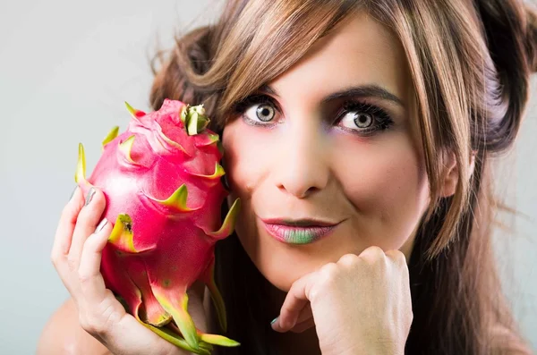 Headshot brunette, dark mystique look and green lipstick, holding up pink pitaya fruit, looking into camera