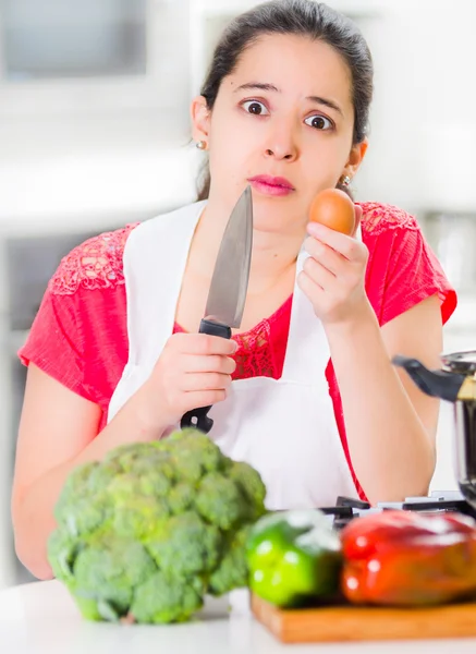 Young woman chef holding up kitchen knife and egg, interacting with facial expressions, vegetables on desk in front