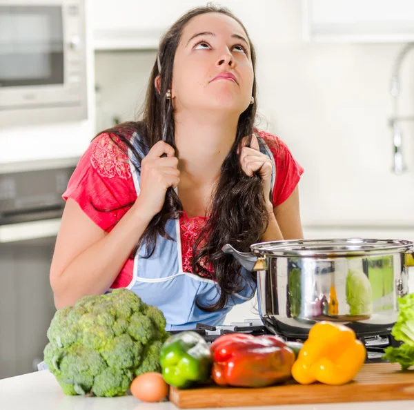 Young woman chef standing over kettle of cooking food, pulling her own hair in frustration and upset facial expression with vegetables on desk
