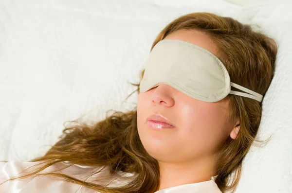 Brunette resting head comfortably on white pillow, blindfold sleeping cover covering eyes