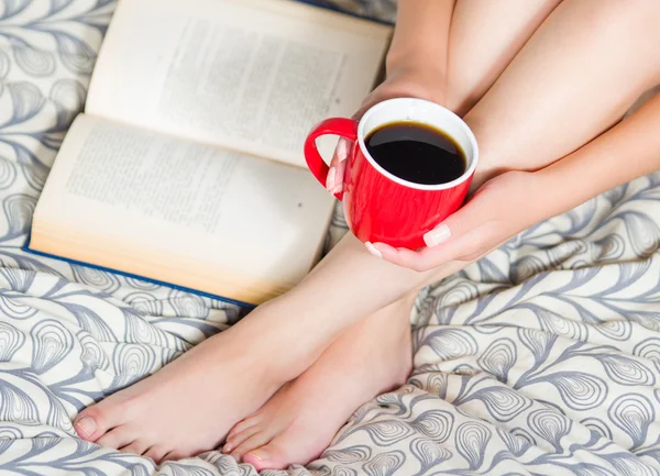 Woman sitting on bed, only legs visible, holding red coffee cup and open book lying next to feet