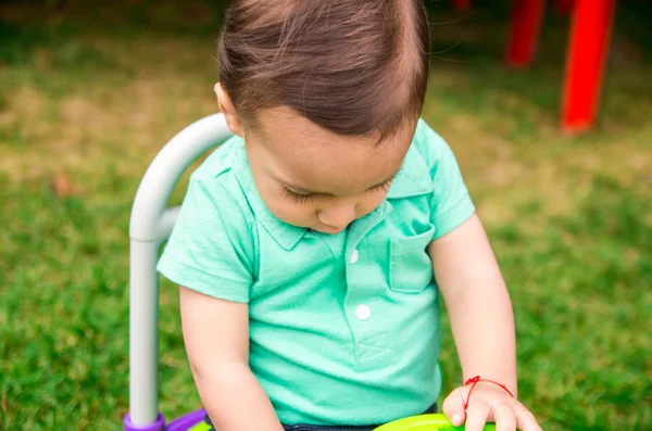 Cute baby boy wearing turquoise t-shirt and combed hair, sitting outside on grassy surface playing around
