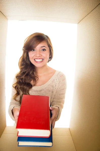 Nice and smiling girl holding two books, red and blue. Inside a cardboard box and white background