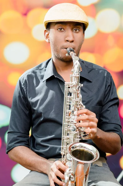 African man wearing sixpence hat and dark shirt playing saxophone, facing camera
