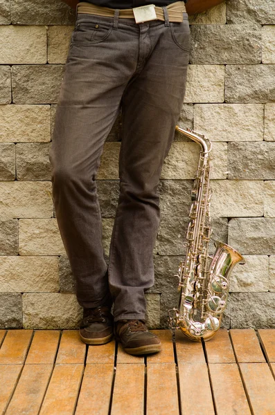 Legs of man standing up against wall with saxophone instrument next to him on floor