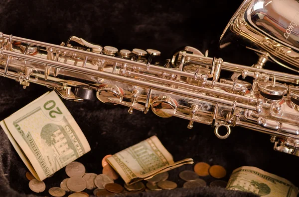Shiny saxophone lying across open instrumental casing with black velvet interior and pile of money inside