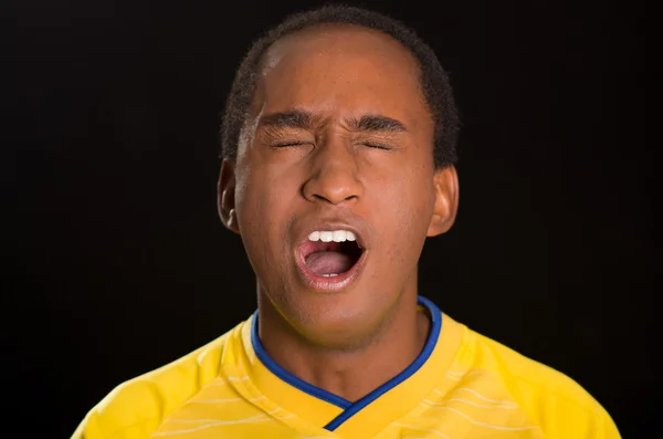 Headshot dark skinned male wearing yellow football shirt in front of black background, eyes closed and mouth open cheering