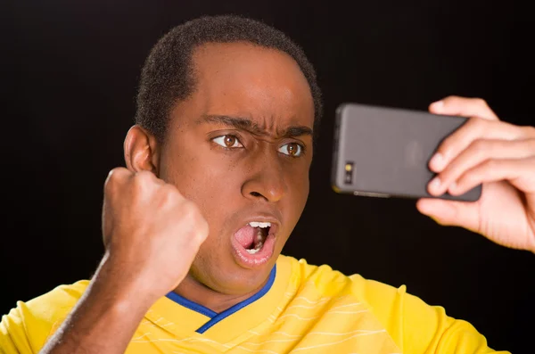 Headshot dark skinned male wearing yellow football shirt in front of black background, holding up mobile phone watching screen and cheering enthusiastically