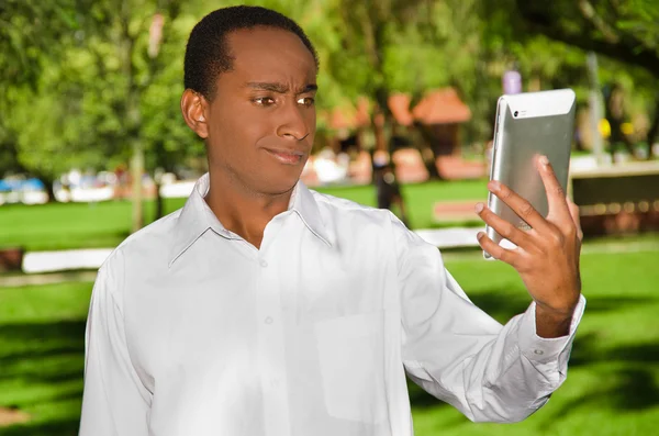 Handsome hispanic black man wearing white shirt in outdoors park area holding up tablet and watching screen as in taking selfie