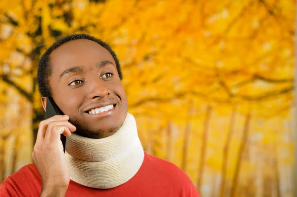 Injured young positive black hispanic male wearing neck brace and talking on phone smiling, yellow abstract background