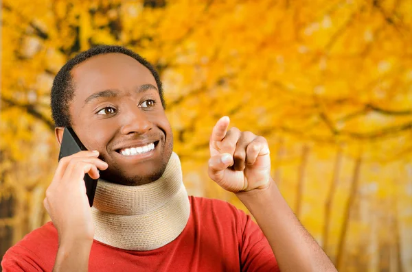 Injured young positive black hispanic male wearing neck brace and talking on phone smiling, yellow abstract background