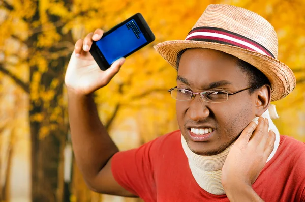 Injured black hispanic male wearing neck brace, glasses and hat, holding cell phone making painful facial expression, yellow abstract background