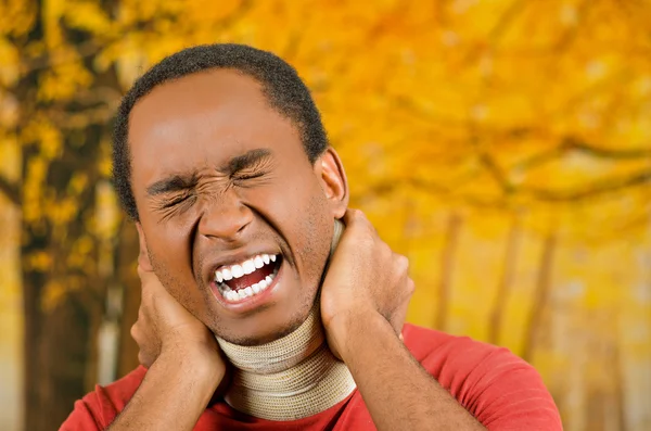 Injured black hispanic male wearing neck brace, holding hands in pain around support making faces of agony, yellow abstract background