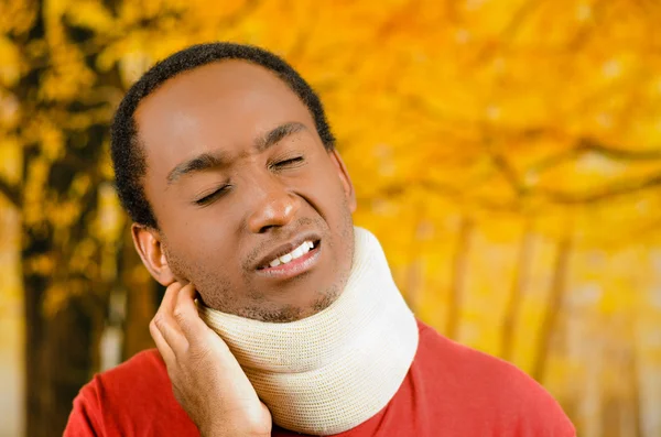Injured black hispanic male wearing neck brace, holding hands in pain around support making faces of agony, yellow abstract background