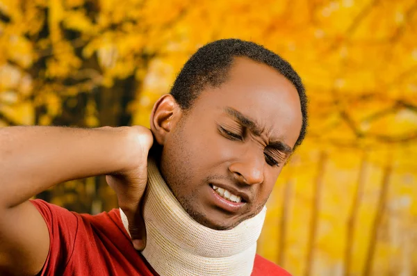 Injured black hispanic male wearing neck brace, holding hands in pain around support making faces of agony, yellow abstract background