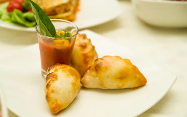 Small tomato soup serving in shot glass with delicious mini empanadas leaning on it, elegant white plate presentation