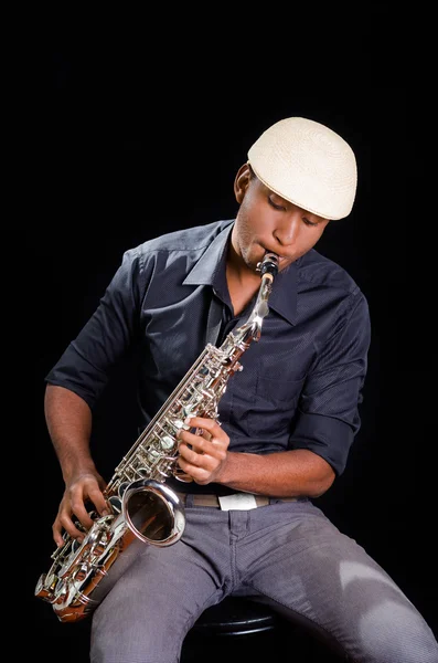 With white hat a black man is sitting and playing his saxophone, dark background, nice music