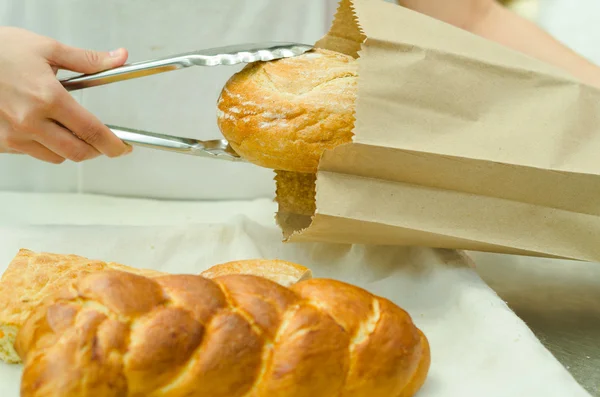 Bakery worker placing loaf of bread inside brown paper bag using large silver tweezers