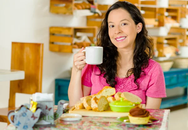 Pretty brunette woman sitting at table inside bakery, holding cup of coffee and smiling happily, bread selection in front