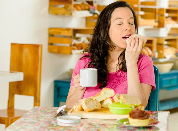 Pretty brunette woman sitting at table inside bakery, holding cup of coffee and biting into bread slice