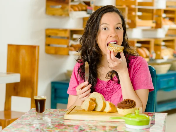Pretty brunette woman wearing pink shirt sitting at table inside bakery, biting into slice of bread