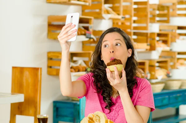 Pretty brunette woman wearing pink shirt sitting by table inside bakery, holding up mobile phone taking a selfie