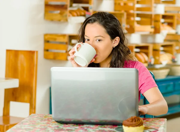 Pretty brunette woman wearing pink shirt sitting by table inside bakery, drinking from coffee mug and looking at laptop screen
