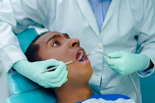 Young hispanic man lying in chair receiving dental treatment with mouth open, dentist hands wearing gloves holding tools working on patients teeth
