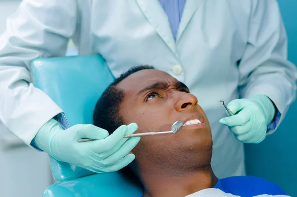 Young hispanic man lying in chair receiving dental treatment with mouth open and upset facial expression, dentist hands wearing gloves holding tools working on patients teeth