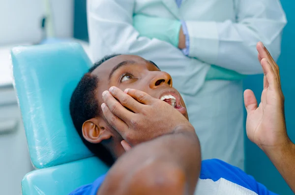 Young hispanic man lying in dental chair holding his mouth and distressed facial expression,