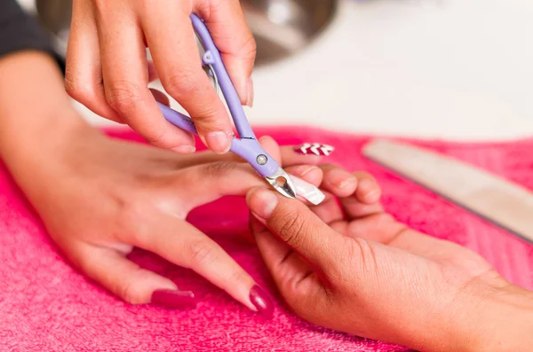 Closeup females hands getting manicure treatment from woman using nail scissors in salon environment, pink towel surface, blurry background products