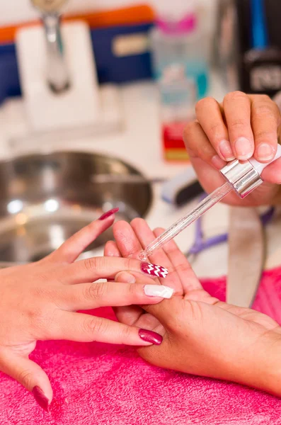 Closeup females hands getting manicure treatment from woman applying transparent liquid in salon environment, pink towel surface, blurry background products