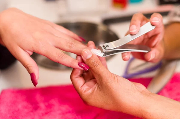 Closeup females hands getting manicure treatment from woman using nailclipper in salon environment, pink towel surface, blurry background products