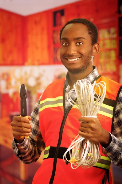 Young electrical worker wearing safety vest, holding cables and cable pliars, smiling with great positive attitude