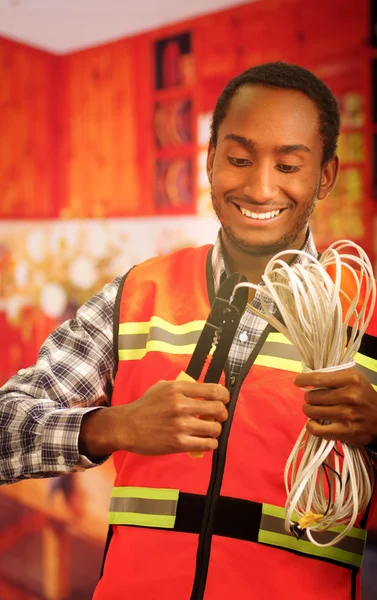 Young electrical worker wearing safety vest, holding cables and cable pliars, smiling with great positive attitude