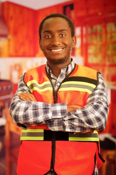 Young carpenter worker wearing square pattern flanel shirt and red safety vest, posing with arms crossed, smiling to camera