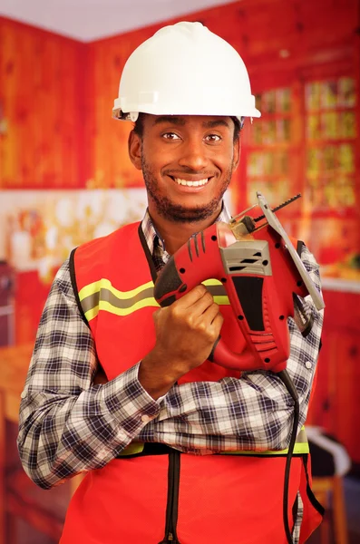 Young engineer carpenter wearing square pattern flanel shirt with red safety vest, holding jigsaw smiling to camera
