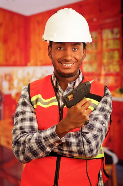 Young engineer carpenter wearing square pattern flanel shirt with red safety vest, holding glue gun smiling to camera
