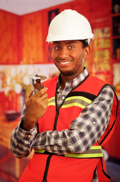 Young engineer carpenter wearing helmet, square pattern flanel shirt with red safety vest, holding small handheld electric saw smiling to camera