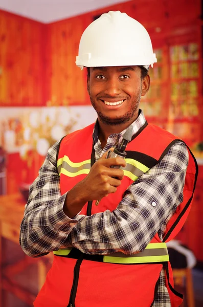 Young engineer carpenter wearing helmet, square pattern flanel shirt with red safety vest, holding small handheld electric polisher tool smiling to camera