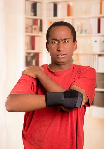 Man in red shirt wearing wrist brace support on right hand posing for camera, holding his shoulder with other arm simulating painful movements