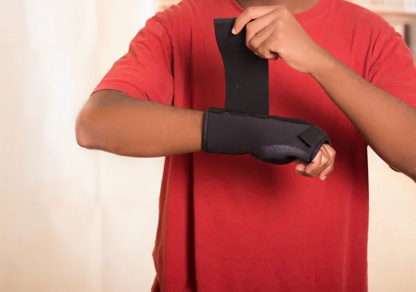 Closeup man in red shirt wearing black wrist brace support on right hand, tightening velcro using other arm