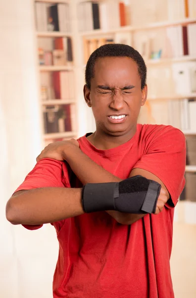 Man in red shirt wearing wrist brace support on right hand posing for camera, holding his shoulder with other arm simulating painful movements