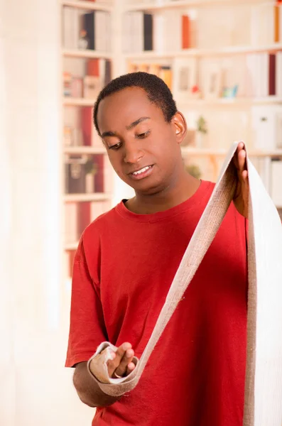 Man in red shirt stretching out bandage preparing wrist support on right hand posing for camera, blurry bookshelves background